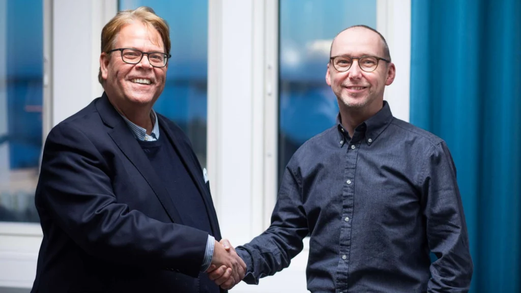 Magnus Johansson och Niclas Eriksson skakar hand framför ett fönster.
